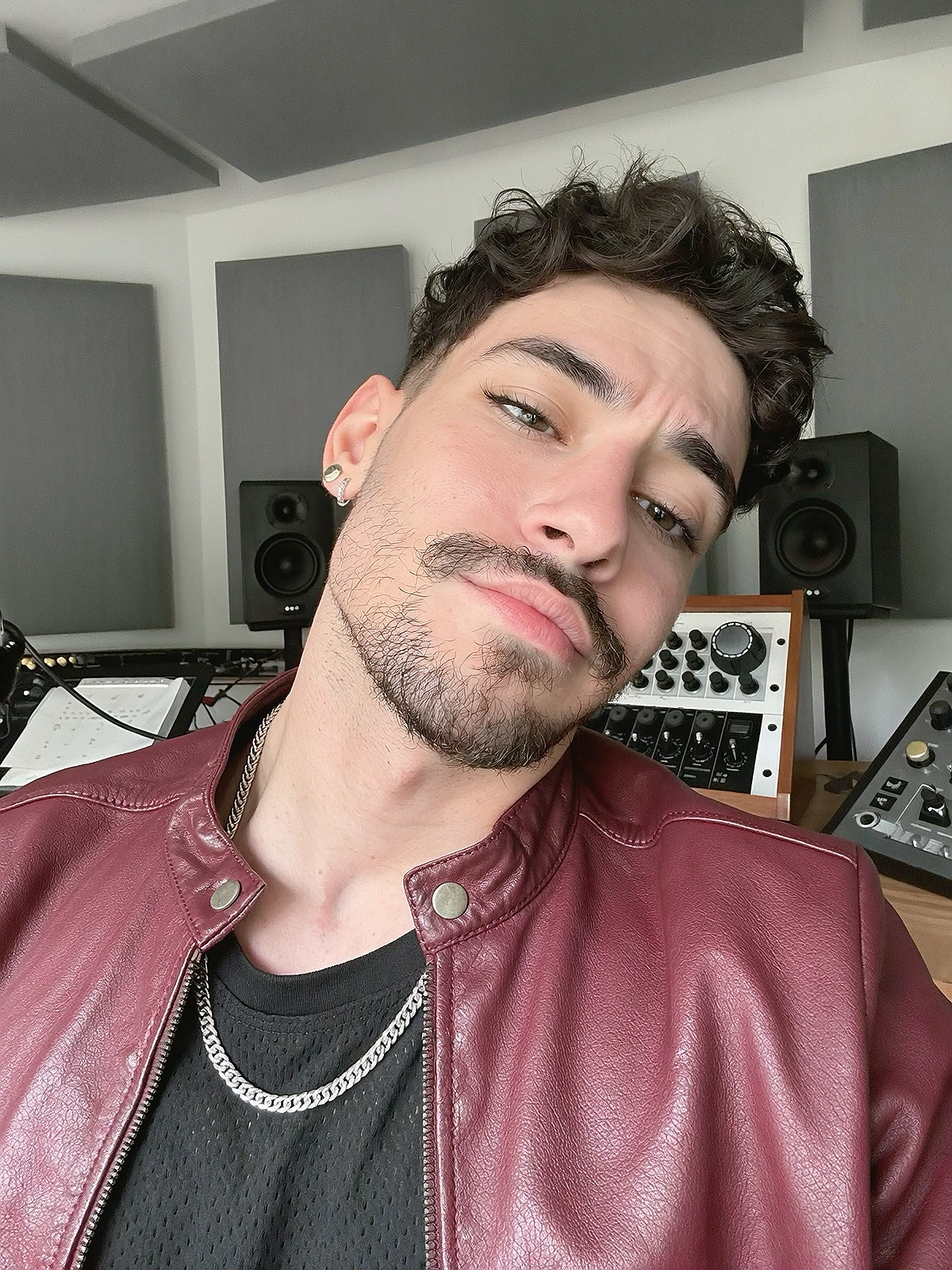 Close shoulder-up candid portrait of a fictional young adult latin / trap artist smiling naturally in their own music recording studio