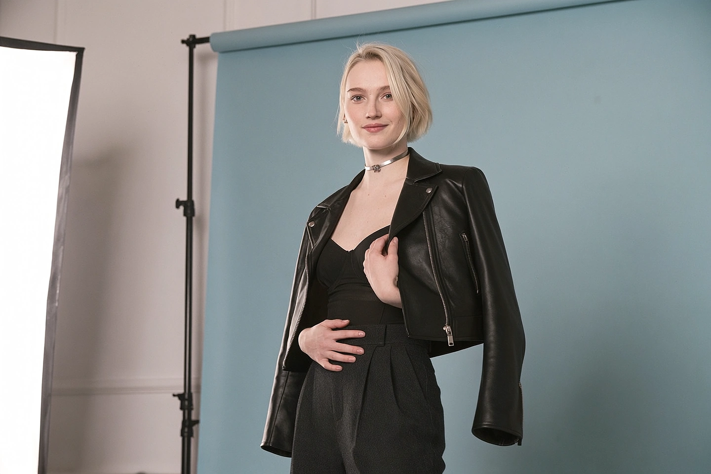 Professional studio photoshoot of a fictional adult pop singer with a blonde bob, black stagewear and a leather jacket against a slate-blue backdrop